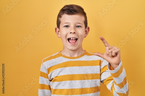 Young caucasian kid standing over yellow background smiling and confident gesturing with hand doing small size sign with fingers looking and the camera. measure concept.