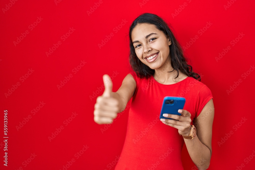 Young brazilian woman using smartphone over red background approving doing positive gesture with hand, thumbs up smiling and happy for success. winner gesture. Young brazilian woman using smartphone over red background approving doing positive gesture with hand, thumbs up smiling and happy for success. winner gesture.
