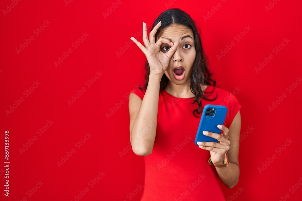Naklejka premium Young brazilian woman using smartphone over red background doing ok gesture shocked with surprised face, eye looking through fingers. unbelieving expression.