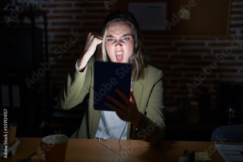 Blonde caucasian woman working at the office at night angry and mad raising fist frustrated and furious while shouting with anger. rage and aggressive concept.
