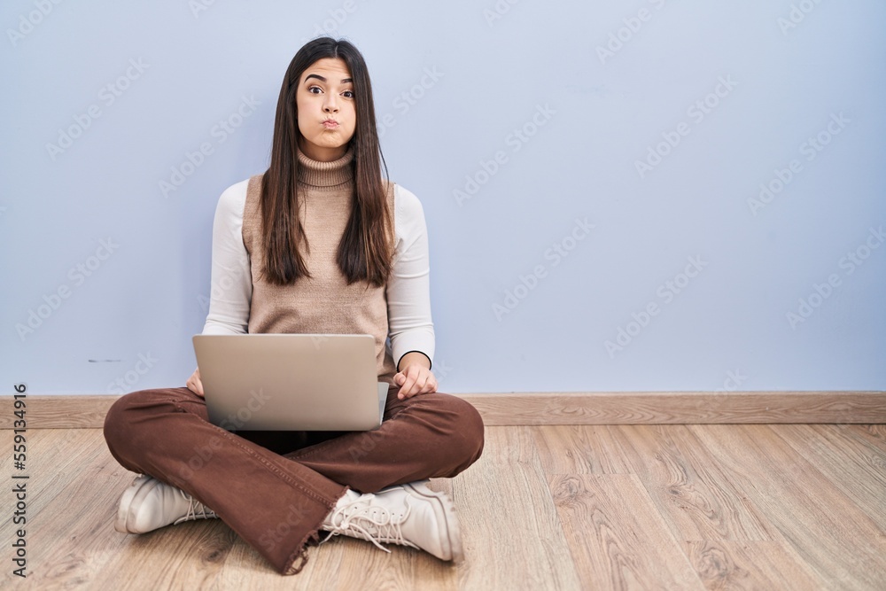 Young brunette woman working using computer laptop sitting on the floor puffing cheeks with funny face. mouth inflated with air, crazy expression. Young brunette woman working using computer laptop sitting on the floor puffing cheeks with funny face. mouth inflated with air, crazy expression.