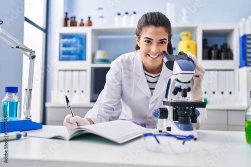 Young beautiful hispanic woman scientist using microscope writing on notebook at laboratory