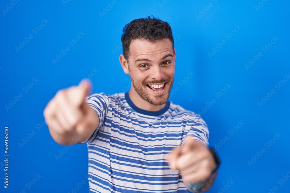 Young hispanic man standing over blue background pointing to you and the camera with fingers, smiling positive and cheerful Young hispanic man standing over blue background pointing to you and the camera with fingers, smiling positive and cheerful