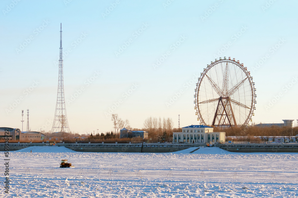 Ferris wheel on the bank of a snowy river against the backdrop of a beautiful sky. Tractor rides on ice on the state border. Winter landscape.