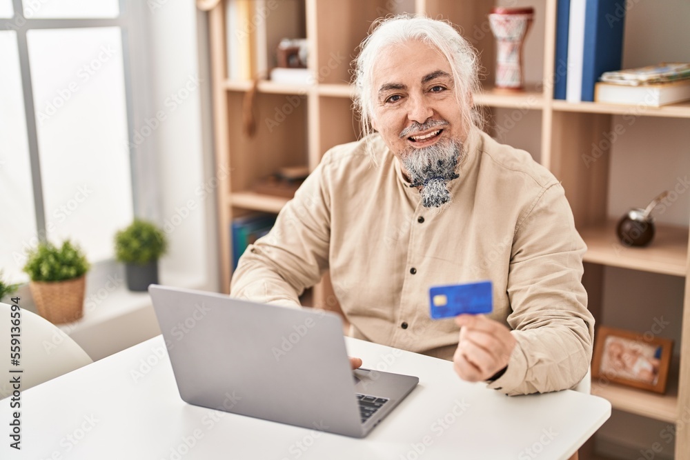 Middle age grey-haired man using laptop and credit card sitting on table at home