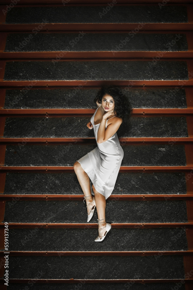 Fototapeta premium Lush hair volume curly hair; A dark-haired girl is dressed in a light elegant dress; a sexy girl lies on the steps of a building; red lipstick on the lips;