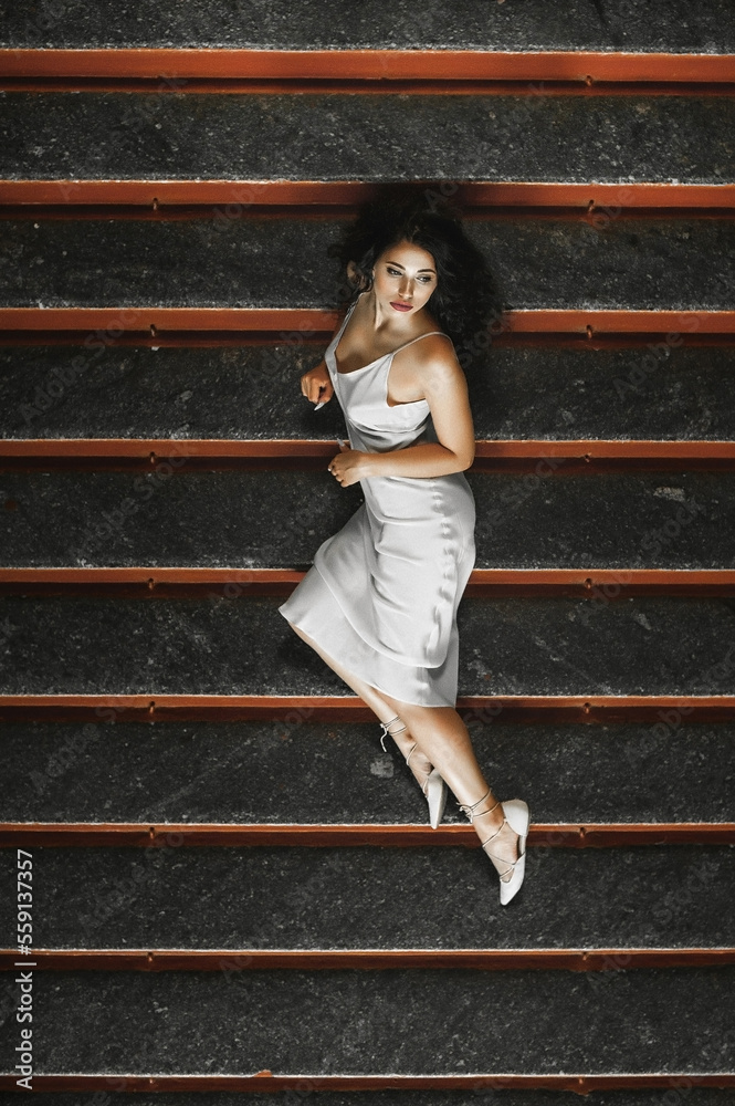 Obraz premium Lush hair volume curly hair; A dark-haired girl is dressed in a light elegant dress; a sexy girl lies on the steps of a building; red lipstick on the lips;