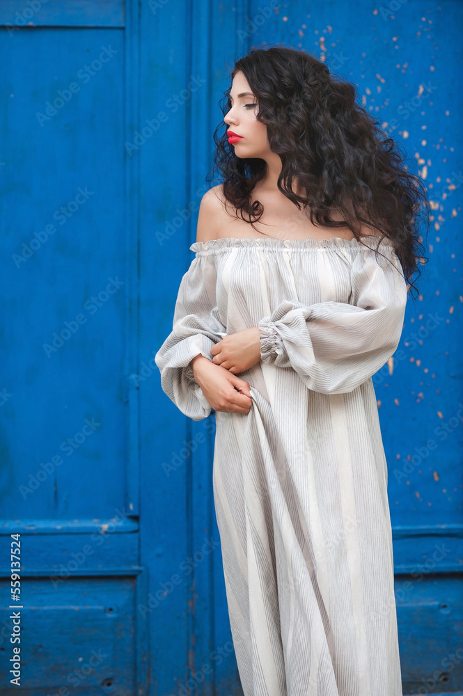 Lush hair volume
 curly hair; A dark-haired girl is wearing a light dress; a beautiful girl poses against the background of a blue door; red lipstick on the lips;