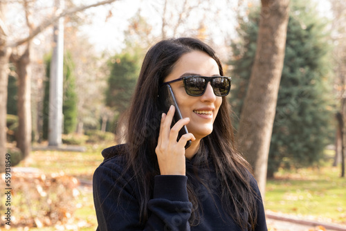 Talking on the phone, millennial brunette woman talking on the phone at park. Happy young girl holding smartphone, answering call. Having pleasant conversation with friend. Walking outdoor on autumn. © Designerant