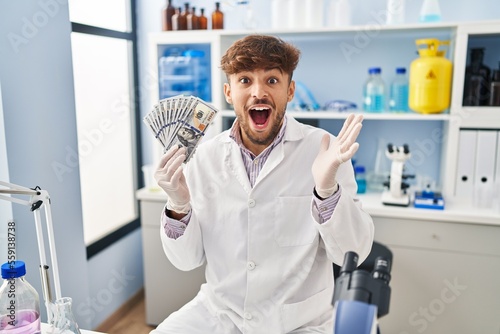 Arab man with beard working at scientist laboratory holding money celebrating victory with happy smile and winner expression with raised hands