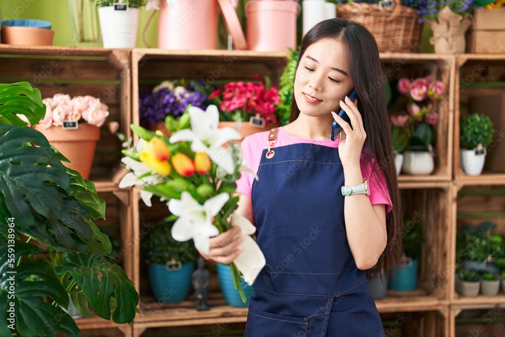 Young chinese woman florist talking on smartphone holding flowers at flower shop