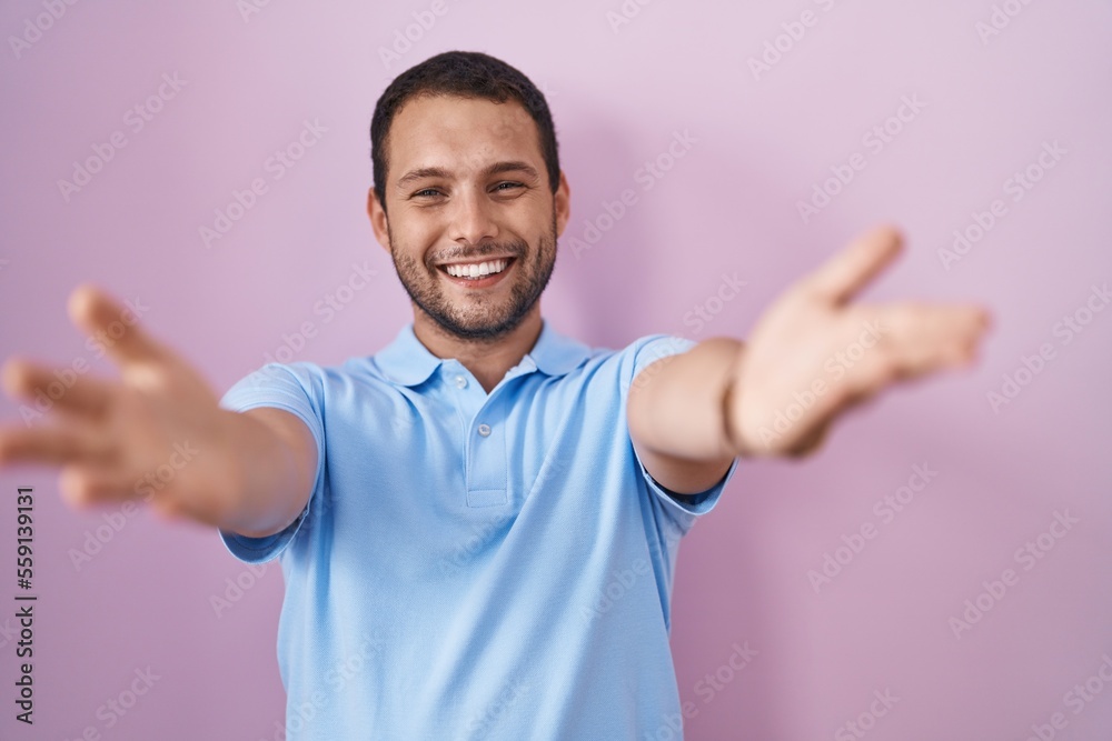 Hispanic man standing over pink background smiling cheerful offering hands giving assistance and acceptance. Hispanic man standing over pink background smiling cheerful offering hands giving assistance and acceptance.