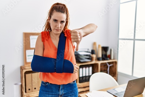 Young redhead woman wearing arm on sling at the office with angry face, negative sign showing dislike with thumbs down, rejection concept © Krakenimages.com