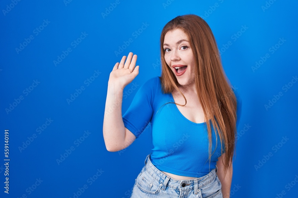 Redhead woman standing over blue background waiving saying hello happy and smiling, friendly welcome gesture Redhead woman standing over blue background waiving saying hello happy and smiling, friendly welcome gesture