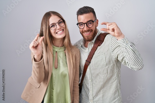 Young couple standing over white background smiling and confident gesturing with hand doing small size sign with fingers looking and the camera. measure concept.
