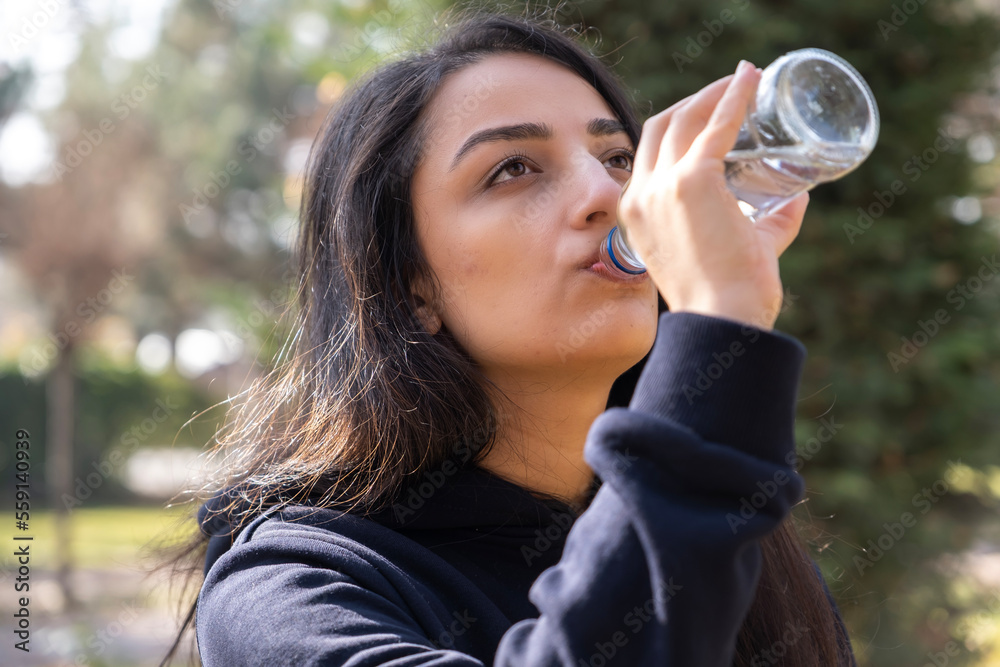 Woman drinking water, lifestyle head shot portrait of woman drinking water. Standing outside, at the park, green background, caucasian lady holding glass bottle enjoying water in autumn. Woman drinking water, lifestyle head shot portrait of woman drinking water. Standing outside, at the park, green background, caucasian lady holding glass bottle enjoying water in autumn.
