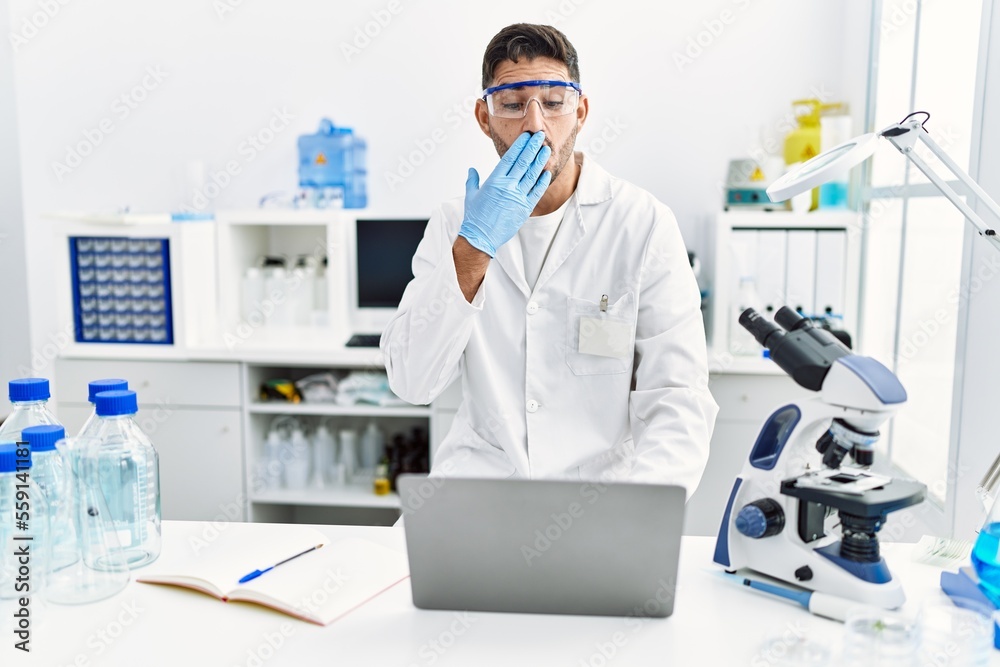 Young hispanic man working at scientist laboratory with laptop covering mouth with hand, shocked and afraid for mistake. surprised expression Young hispanic man working at scientist laboratory with laptop covering mouth with hand, shocked and afraid for mistake. surprised expression