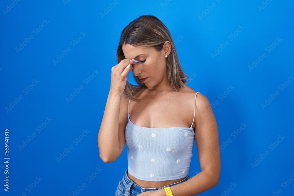 Young woman standing over blue background tired rubbing nose and eyes feeling fatigue and headache. stress and frustration concept. Young woman standing over blue background tired rubbing nose and eyes feeling fatigue and headache. stress and frustration concept.