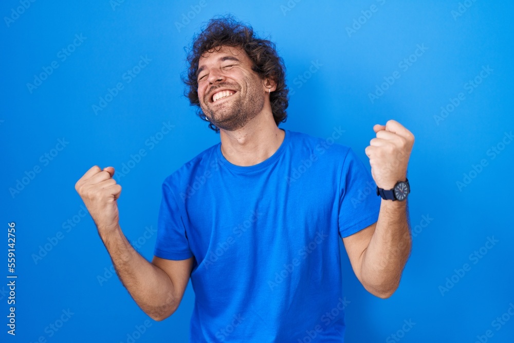 Hispanic young man standing over blue background celebrating surprised and amazed for success with arms raised and eyes closed. winner concept. Hispanic young man standing over blue background celebrating surprised and amazed for success with arms raised and eyes closed. winner concept.