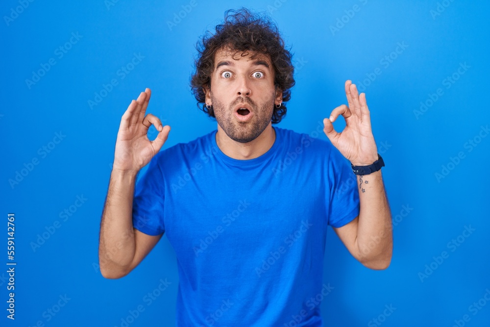 Hispanic young man standing over blue background looking surprised and shocked doing ok approval symbol with fingers. crazy expression Hispanic young man standing over blue background looking surprised and shocked doing ok approval symbol with fingers. crazy expression