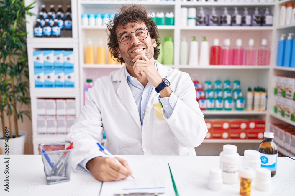 Hispanic young man working at pharmacy drugstore with hand on chin thinking about question, pensive expression. smiling and thoughtful face. doubt concept.