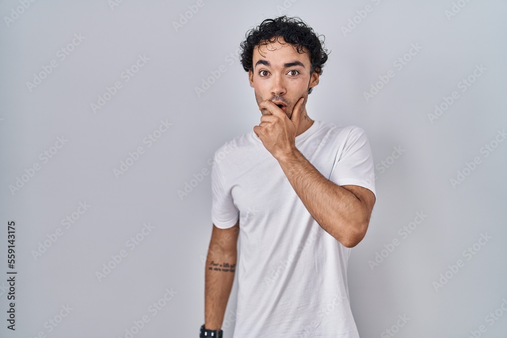 Hispanic man standing over isolated background looking fascinated with disbelief, surprise and amazed expression with hands on chin Hispanic man standing over isolated background looking fascinated with disbelief, surprise and amazed expression with hands on chin