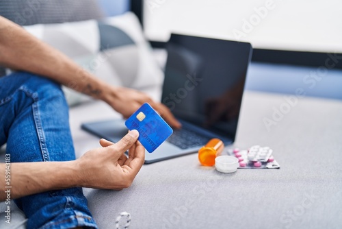 Young hispanic man using laptop and credit card sitting on sofa at home