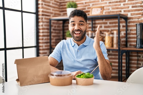 Hispanic man with beard eating delivery salad celebrating victory with happy smile and winner expression with raised hands