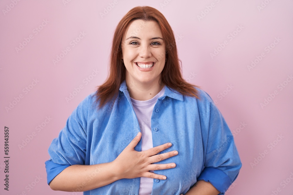 Young hispanic woman with red hair standing over pink background smiling and laughing hard out loud because funny crazy joke with hands on body.