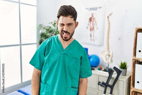 Young man with beard working at pain recovery clinic winking looking at the camera with sexy expression, cheerful and happy face.