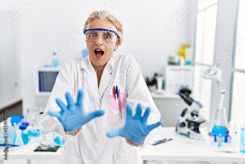 Middle age blonde woman working at scientist laboratory afraid and terrified with fear expression stop gesture with hands, shouting in shock. panic concept.