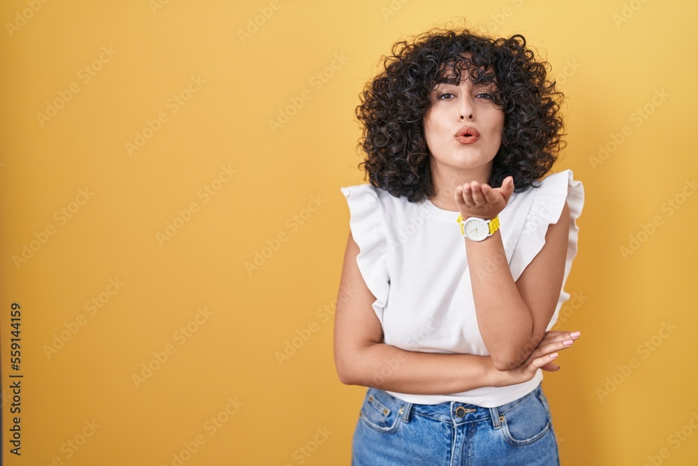 Young middle east woman standing over yellow background looking at the camera blowing a kiss with hand on air being lovely and sexy. love expression. Young middle east woman standing over yellow background looking at the camera blowing a kiss with hand on air being lovely and sexy. love expression.