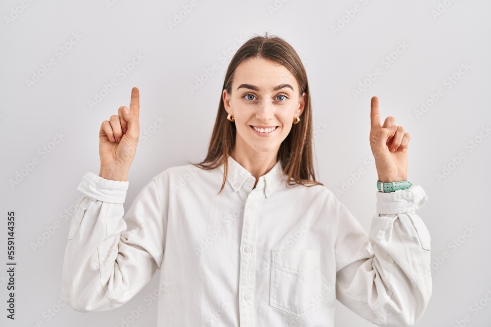 Young caucasian woman standing over isolated background smiling amazed and surprised and pointing up with fingers and raised arms. Young caucasian woman standing over isolated background smiling amazed and surprised and pointing up with fingers and raised arms.