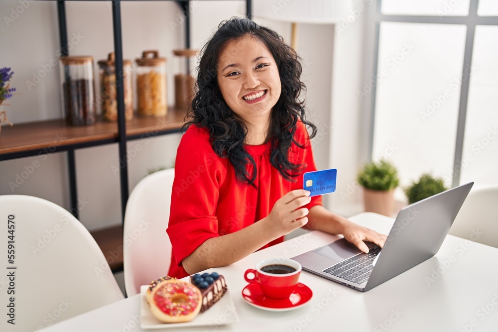 Young chinese woman having breakfast using laptop and credit card at home
