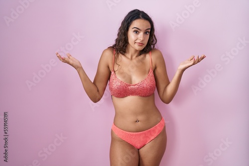 Young hispanic woman wearing lingerie over pink background clueless and confused expression with arms and hands raised. doubt concept.