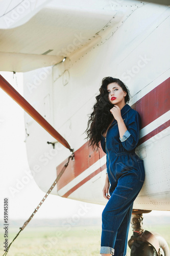 Magnificent volume curly hair; A dark-haired girl is wearing denim overalls; a sexy girl poses against the background of an airplane; red lipstick on the lips;