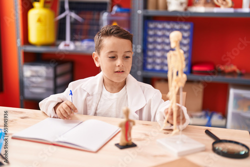 Adorable hispanic toddler student writing on notebook looking skeleton at classroom