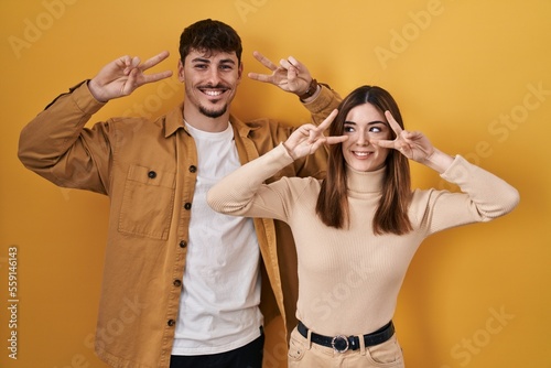 Young hispanic couple standing over yellow background doing peace symbol with fingers over face, smiling cheerful showing victory