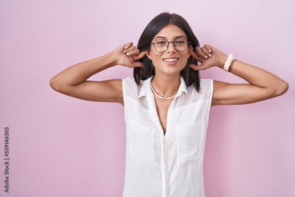 Brunette young woman standing over pink background wearing glasses smiling pulling ears with fingers, funny gesture. audition problem Brunette young woman standing over pink background wearing glasses smiling pulling ears with fingers, funny gesture. audition problem