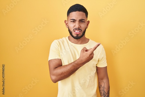 Young hispanic man standing over yellow background pointing aside worried and nervous with forefinger, concerned and surprised expression