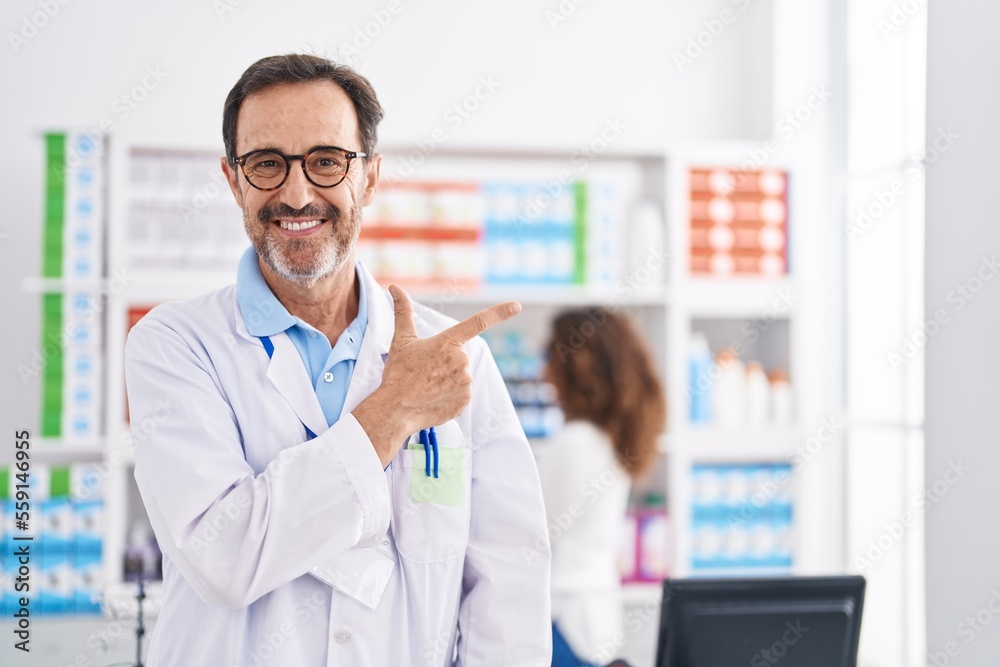 Middle age hispanic man working at pharmacy drugstore smiling cheerful pointing with hand and finger up to the side