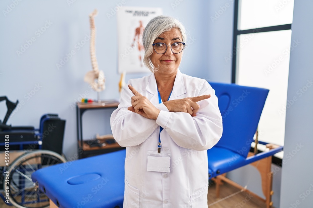 Middle age woman with grey hair working at pain recovery clinic pointing to both sides with fingers, different direction disagree