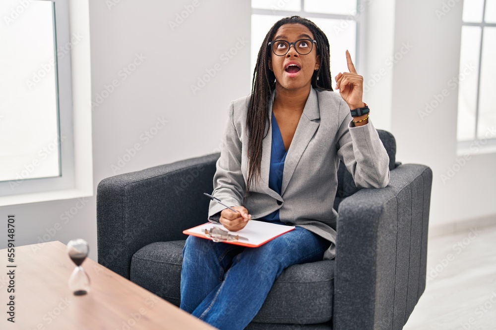 Young african american with braids working at consultation office amazed and surprised looking up and pointing with fingers and raised arms. Young african american with braids working at consultation office amazed and surprised looking up and pointing with fingers and raised arms.