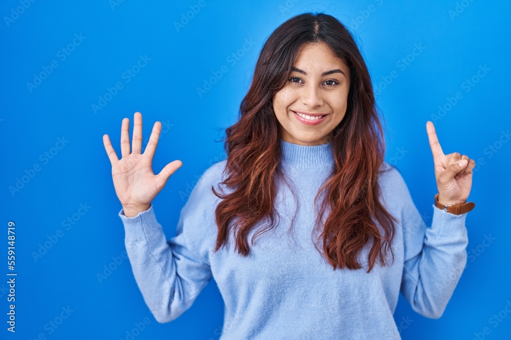 Hispanic young woman standing over blue background showing and pointing up with fingers number six while smiling confident and happy. Hispanic young woman standing over blue background showing and pointing up with fingers number six while smiling confident and happy.