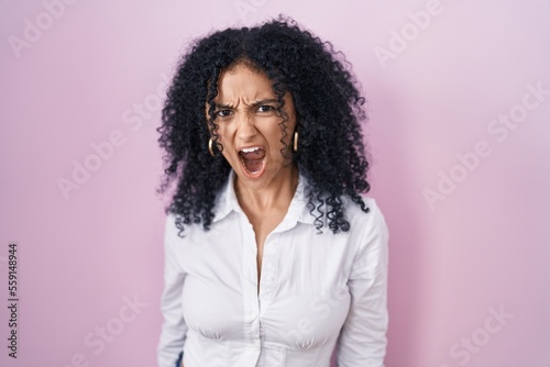 Hispanic woman with curly hair standing over pink background angry and mad screaming frustrated and furious, shouting with anger. rage and aggressive concept.