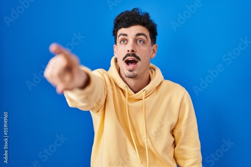 Hispanic man standing over blue background pointing with finger surprised ahead, open mouth amazed expression, something on the front
