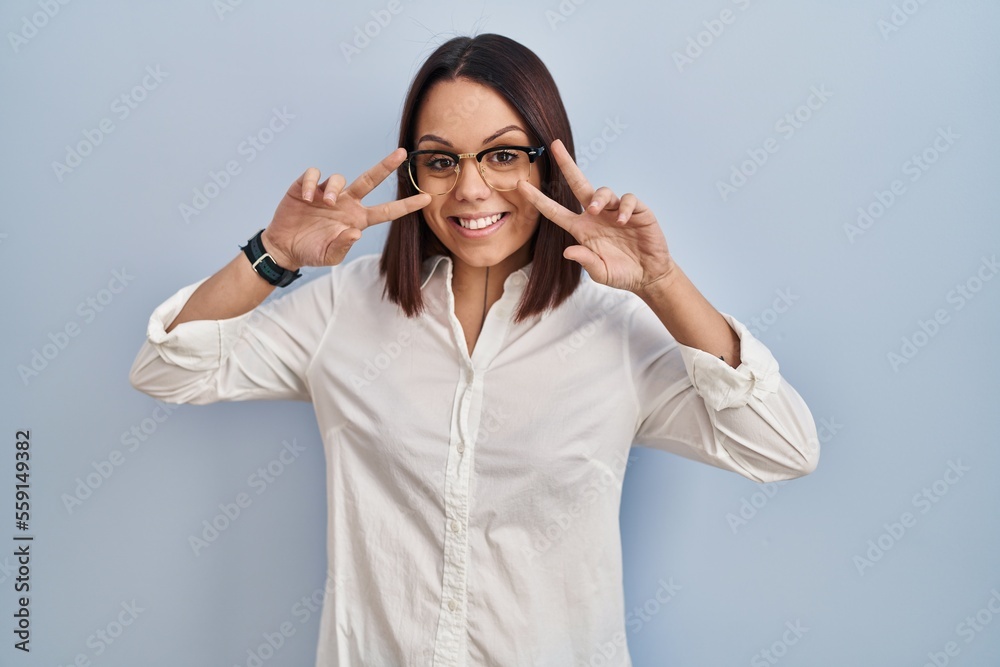 Young hispanic woman standing over white background doing peace symbol with fingers over face, smiling cheerful showing victory Young hispanic woman standing over white background doing peace symbol with fingers over face, smiling cheerful showing victory