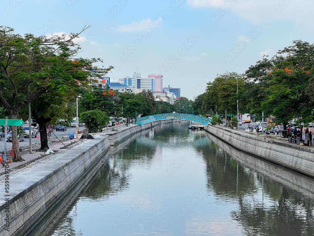 Bangkok, Khlong Phadung Krung Kasem canal. View to the beautiful small walking bridge and hight buildings in the distance. Reflection of blue sky, white clouds and green trees on the surface of water.