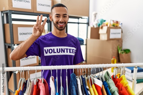 African american man wearing volunteer t shirt at donations stand showing and pointing up with fingers number three while smiling confident and happy.