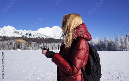A young woman holds a cup of tea against the background of mountains in winter. Strbske Pleso Lake, Slovakia